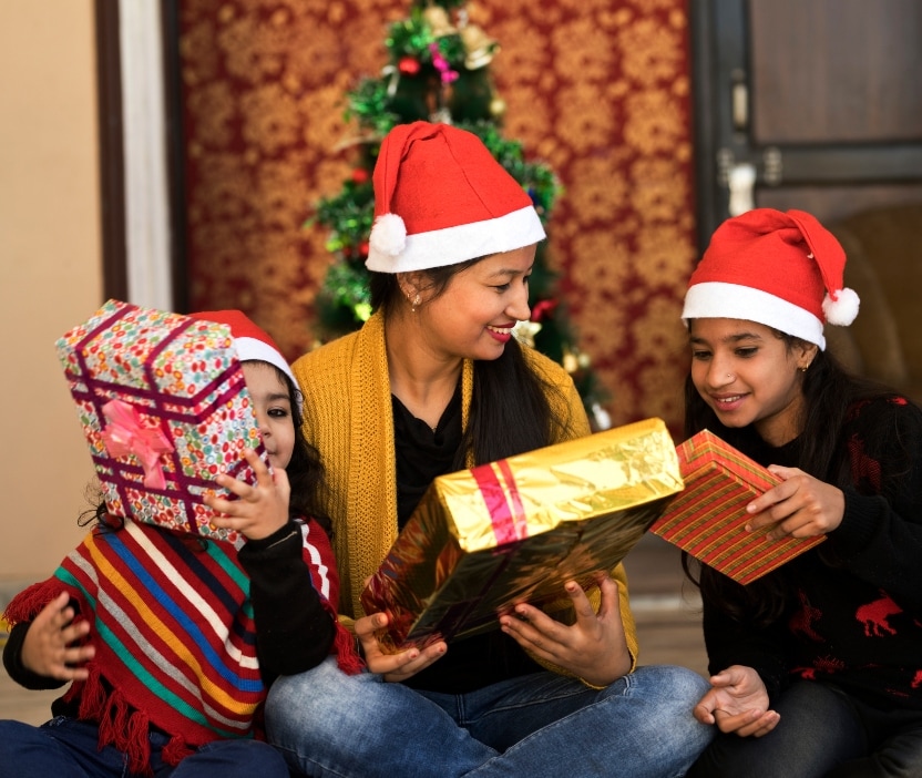 A family of three sits in front of a Christmas tree wearing Santa hats and holding gift boxes.