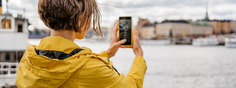 A woman snaps a picture of a landmark on her travels but is unaware that someone is watching her through the phone camera.