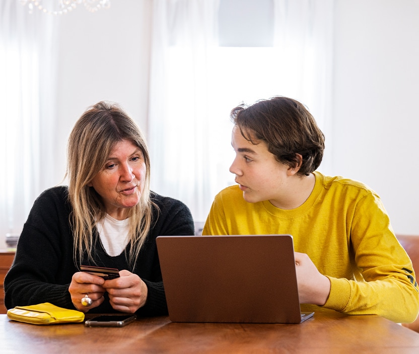 A woman sits at a table with her son looking at a laptop where they are encountering a scammer using loss aversion to influencer them to fall for a scam. 