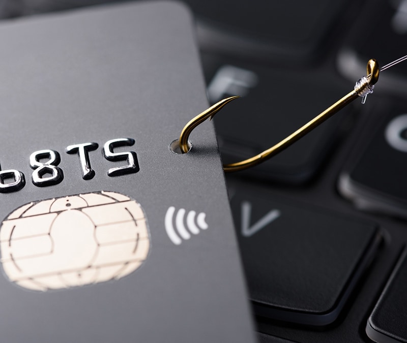 A credit card on top of a keyboard with a fishing hook piercing through it, representing phishing scams.