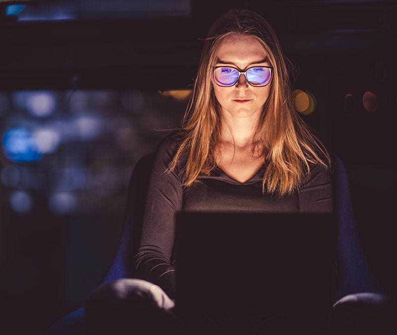 A photograph of a woman using her laptop in a darkened room.