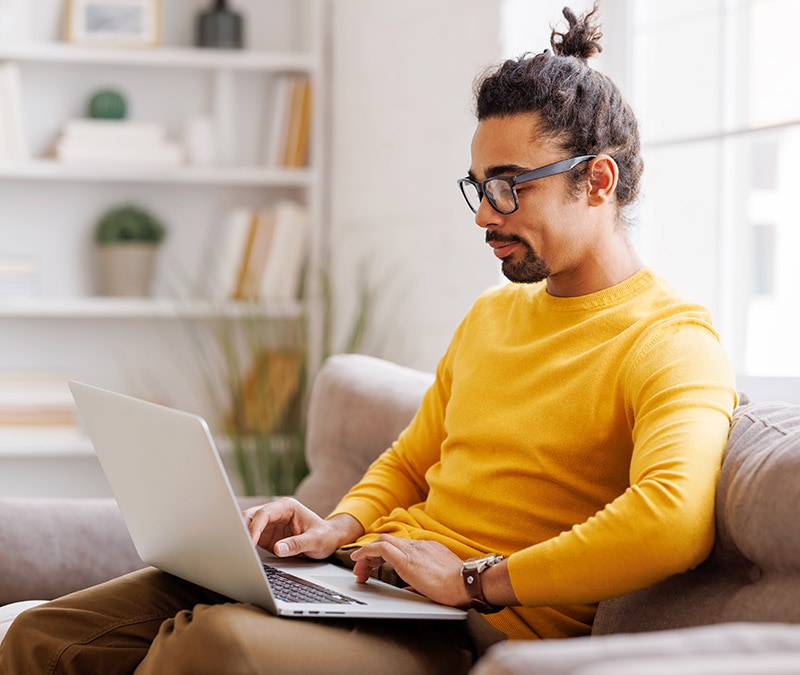 A man in glasses and a yellow shirt sitting on a couch with a laptop, engrossed in his work.