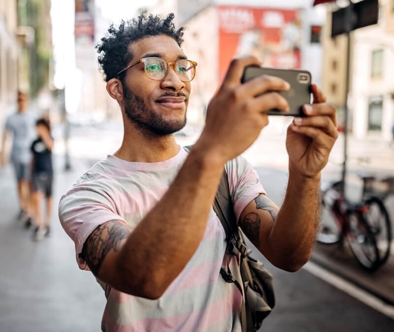 A man walking in a city stops to take a photo on his smartphone.