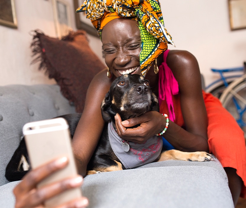 A woman checks looks on her phone while laying on a sofa with her dog, and she is learning about if pet tech can be hacked.