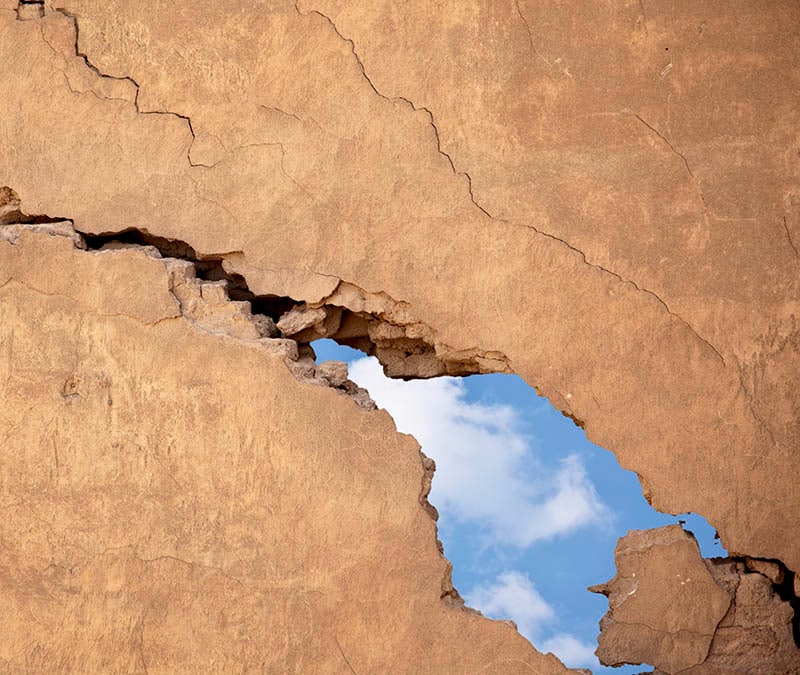 A large crack in a sheet of rock offers a glimpse of a blue sky with clouds.