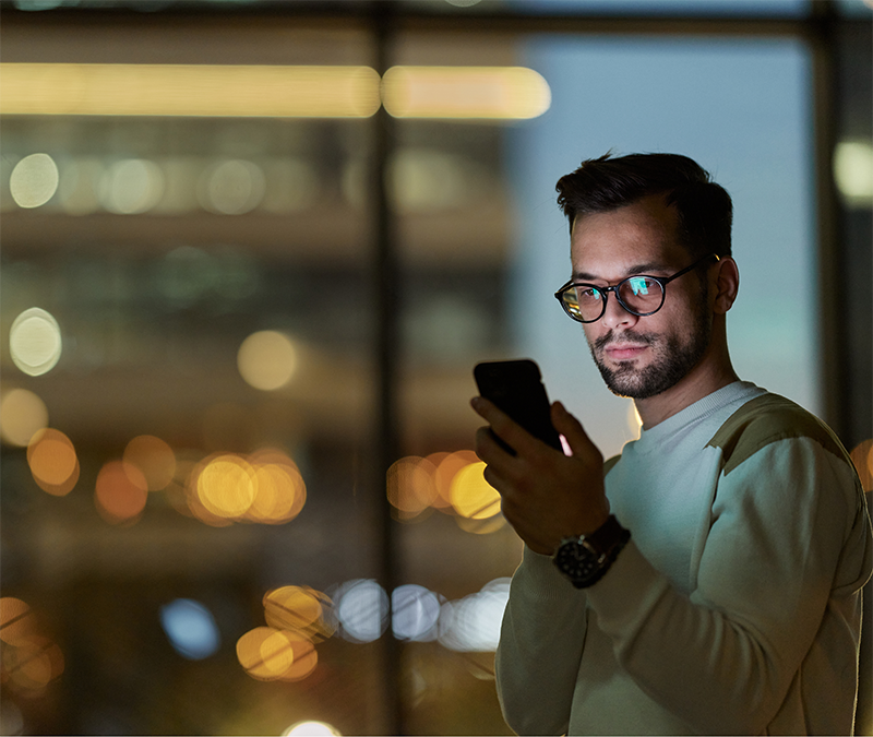 A young man wearing glasses and a light sweater stands indoors at night, looking at his smartphone with a focused expression. Behind him, large windows reveal an urban cityscape with blurred lights and buildings.
