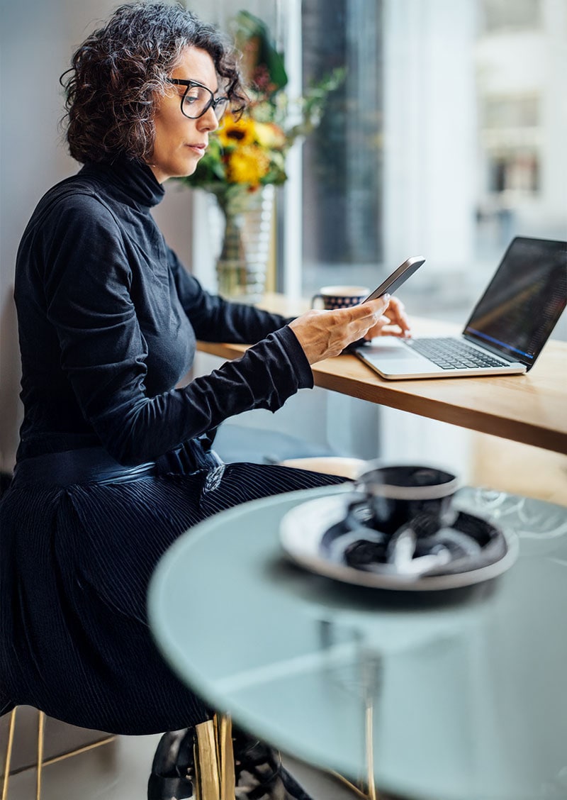 A woman with glasses sits at a table in front of a laptop while looking at her phone.