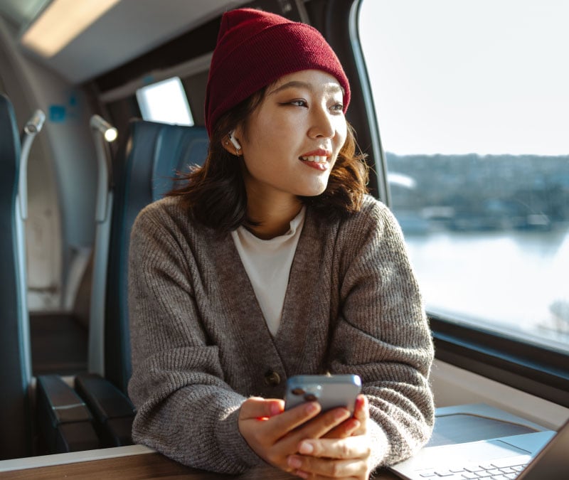 A woman traveling on a train holds her phone while sitting at a table with an open laptop in front of her.