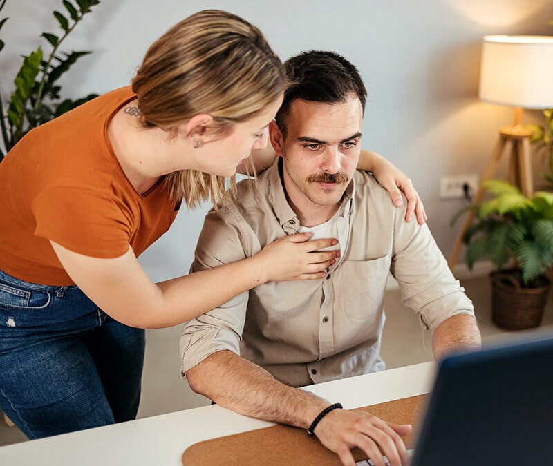 A man sits at a laptop with a somber, stressed expression as a woman stands beside him, wrapping her arms around his shoulders in a supportive embrace.