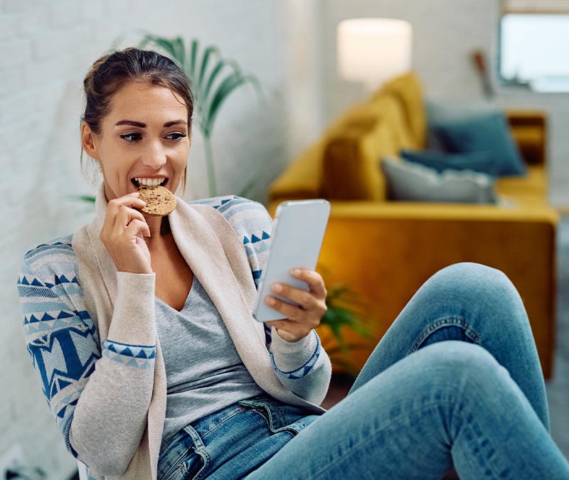 A woman eating a cookie and scrolling on her phone, symbolizing the use of third-party web cookies.
