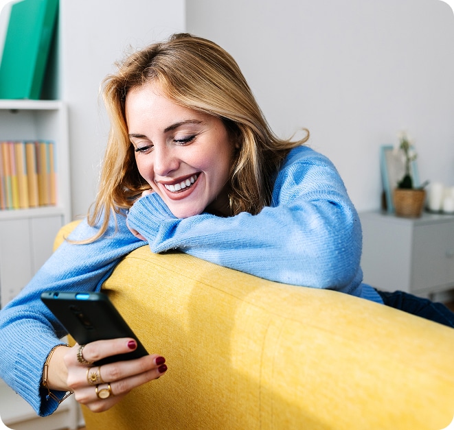 A woman sitting on a couch, looking at her phone, and smiling at a conversation with an AI chatbot. 