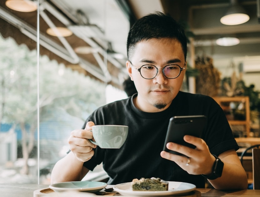 Man sitting in a coffee shop holding a mug of coffee and looking at his phone.