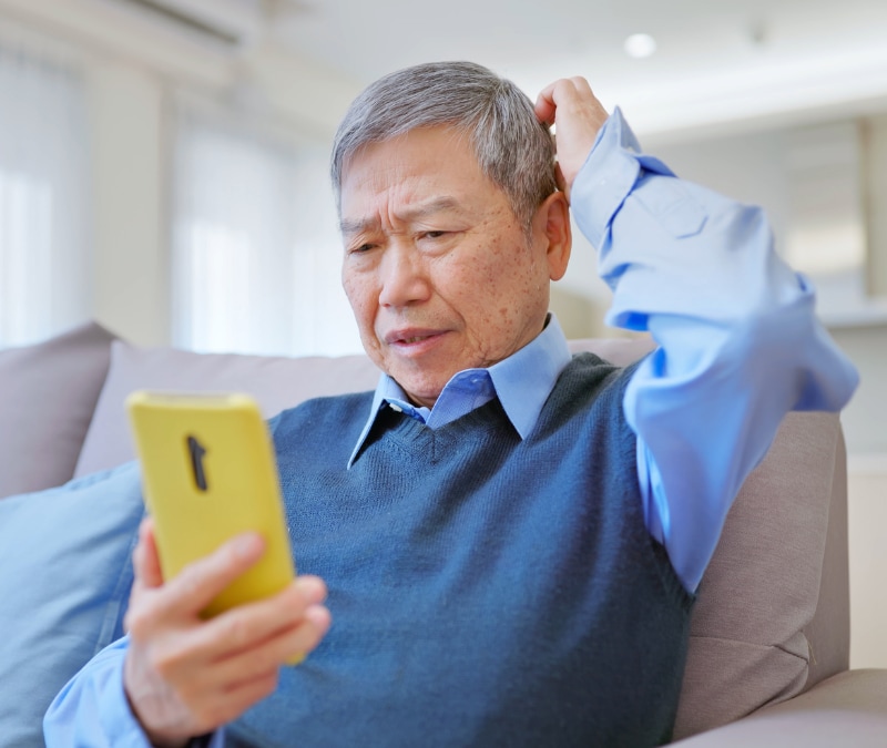 A senior man sits on a couch with his mobile device confused because he was the target of a phone scam. 
