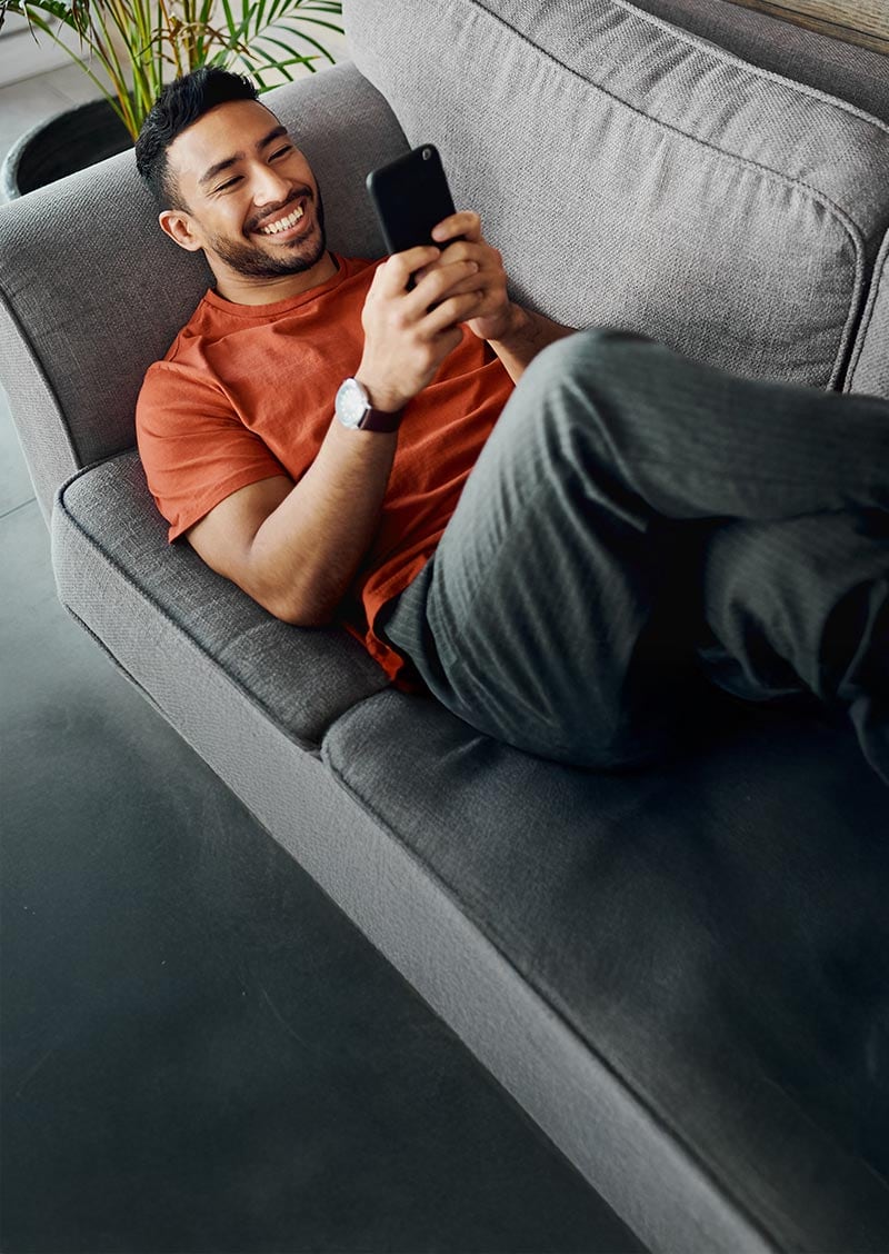 A man lying on a couch and smiling while using a dating app on his phone.