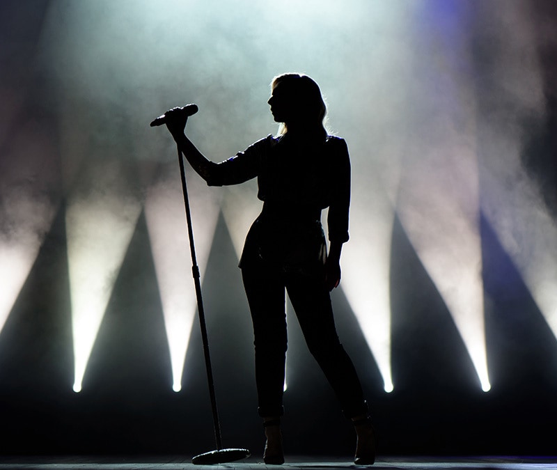 A silhouette of a lady singing at a concert venue.