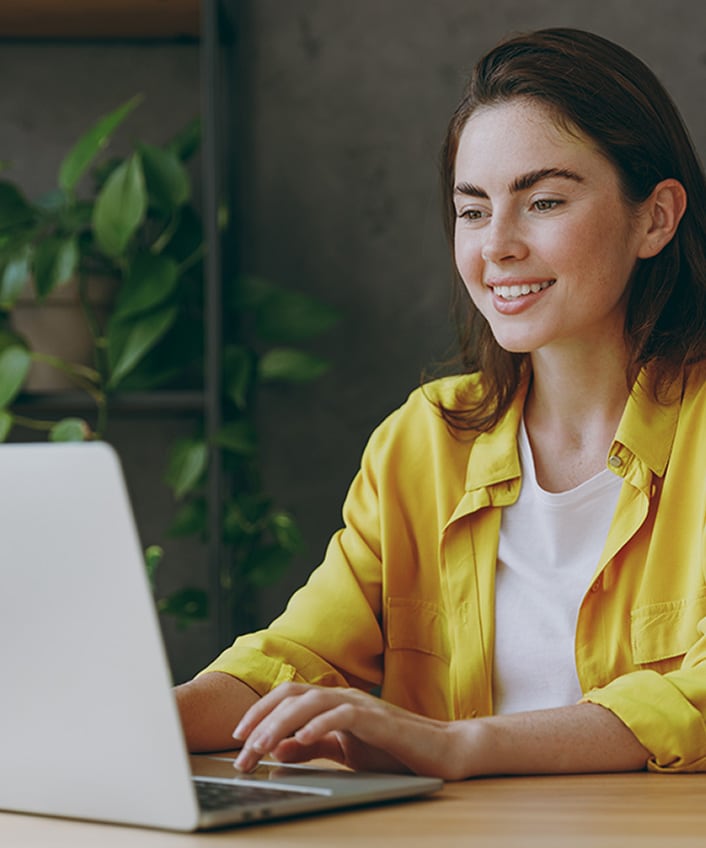 A woman with a yellow shirt looking at her laptop learning about the benefits of a VPN.
