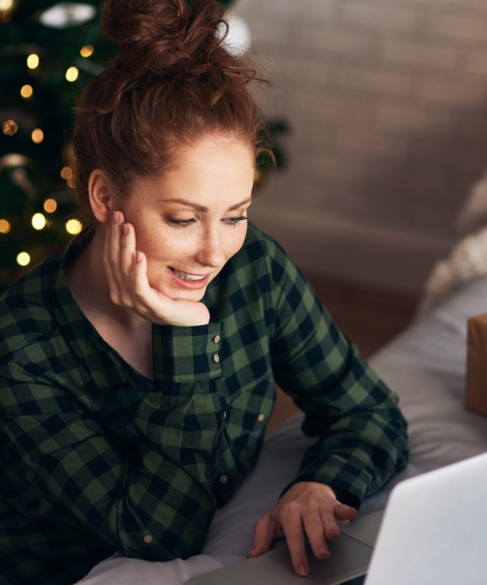 A woman sits at her laptop with a Christmas tree behind her and presents on the bed next to her.