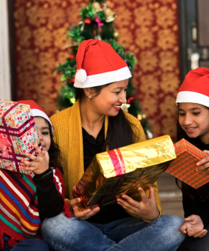 A family of three sits in front of a Christmas tree wearing Santa hats and holding gift boxes.