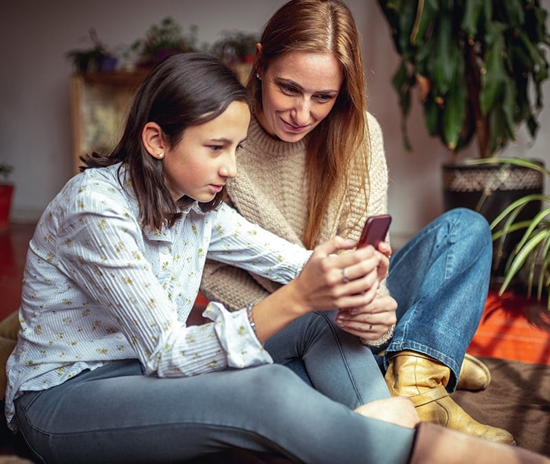 A mother sits next to her daughter, ensuring the iPhone parental controls on her daughter's phone are working properly.