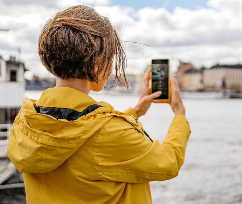 A woman snaps a picture but is unaware that someone is watching her through the phone camera.