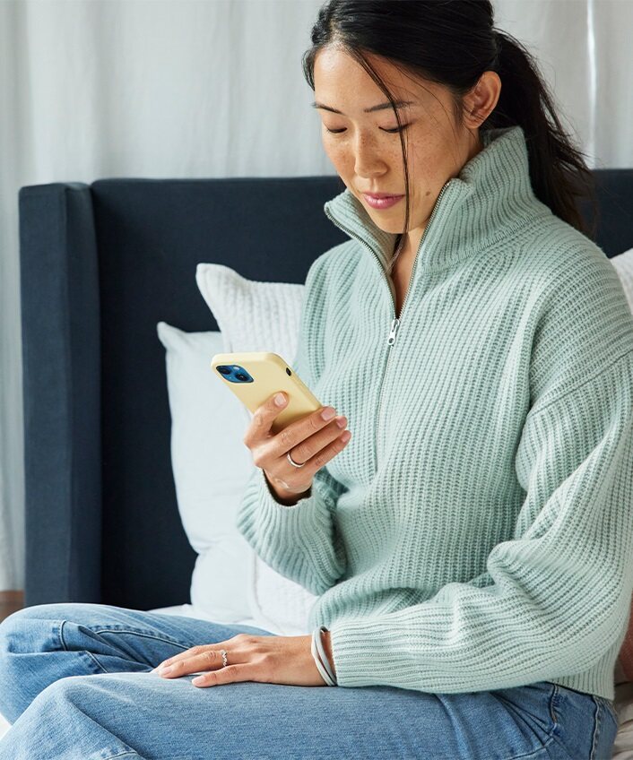Woman sitting on the side of her bed looking at her phone.