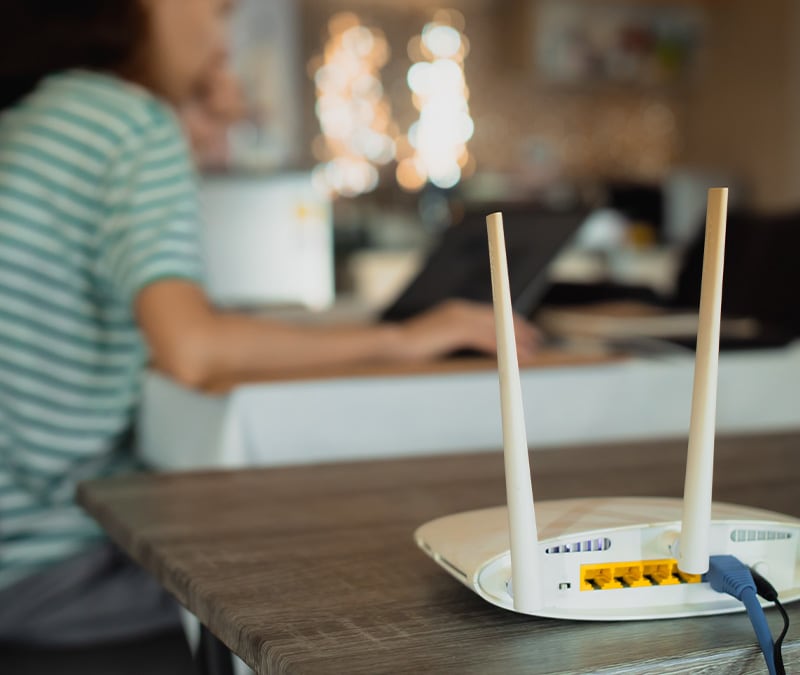 A router on a table with a person browsing on a laptop in the background, possibly targeted by an IP spoofing attack.