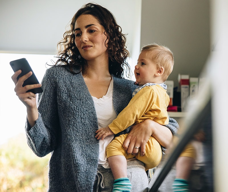 A woman holding her baby in one arm and using a smartphone in the other hand.