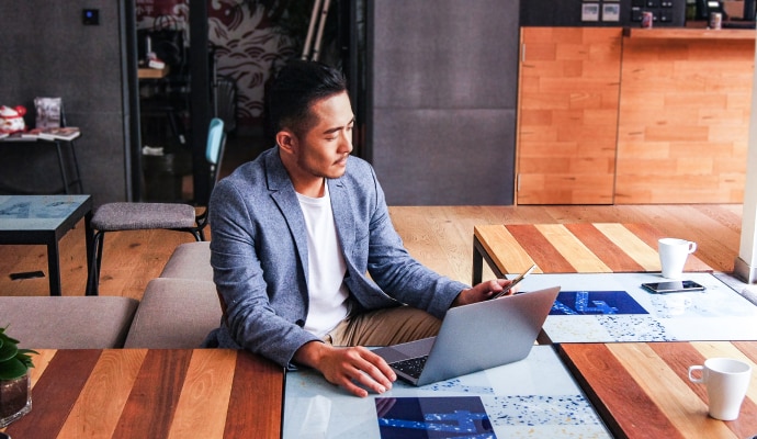 A man looking at a mobile phone while sitting in front of a laptop