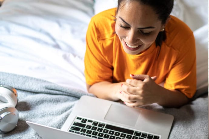 A woman laying on bed smiling using a Laptop.